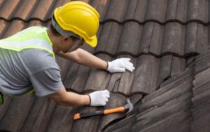 a roofer wearing a yellow hard hat and safety vest inspects damaged concrete roof tiles, with a hammer resting on the roof surface nearby.