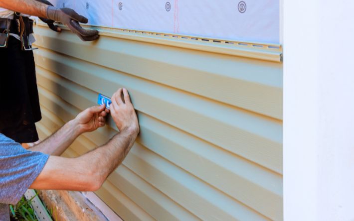 A close-up of a worker using a small blue spirit level to ensure a row of light green vinyl siding is straight during installation on a house wall.