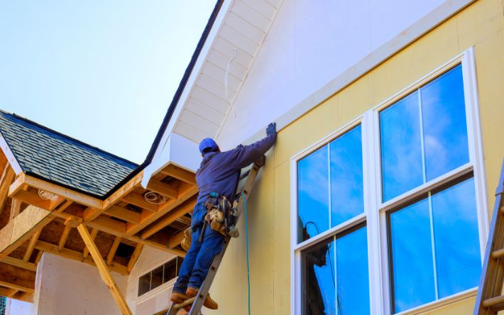 a construction worker wearing a blue cap stands on a ladder to install white fascia trim on the roofline of a two-story house with yellow exterior walls and large windows.